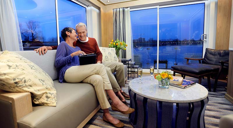 A couple sitting on a couch in a Viking River Ship Stateroom with the night-time city views out the window.